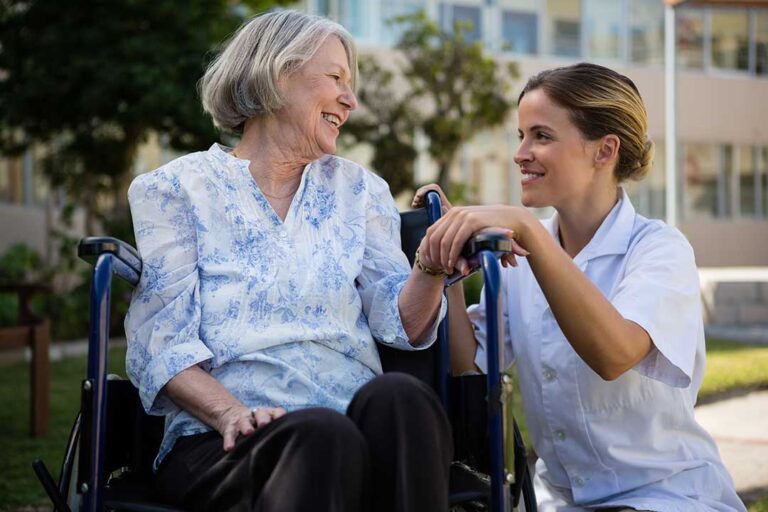 Two assisted living staff members help an older resident with morning exercise in a sunny common room.