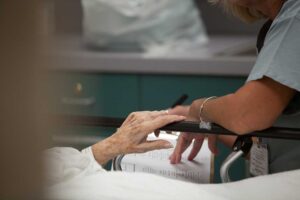 Older adult using a hallway handrail while staff member checks a well-lit assisted living corridor.